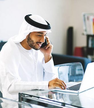 Traditionally dressed Middle Eastern man is talking on phone while surfing the net at home. Image contains some copy space.
